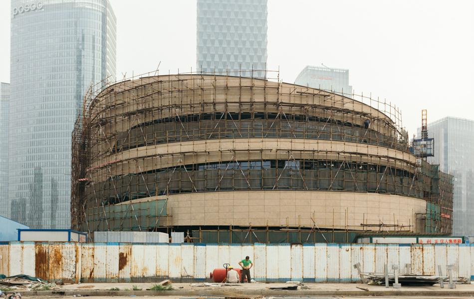 01 斗獸場(chǎng) Colosseum  2016年，北京阿里巴巴大廈旁邊一座建設(shè)中的環(huán)形建筑，兩個(gè)建筑工人正在休息。其中一個(gè)擼起衣服露出肚皮，好似是羅馬斗獸場(chǎng)里的勇士一樣，面對(duì)著背后的這個(gè)龐然大物。_